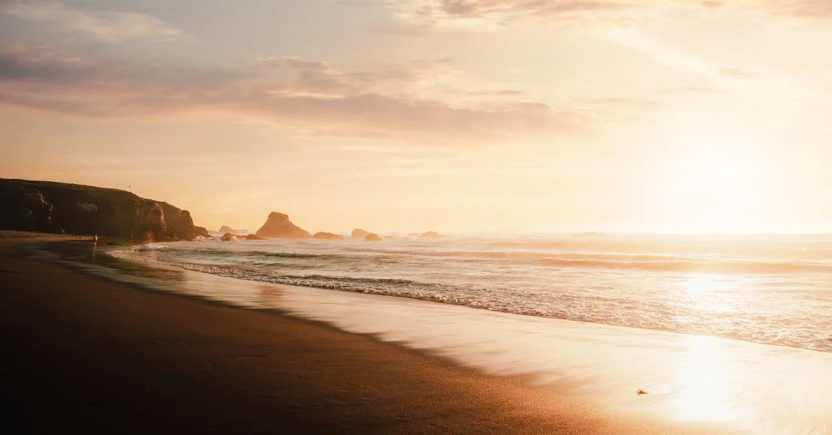 Picturesque view of evening sky covered with light pink clouds above sandy beach and mountains on rocky shore of sea in sunlight