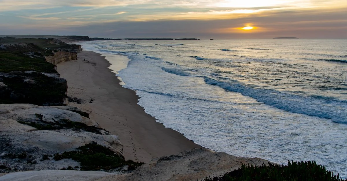 Stunning sunset over a tranquil beach with rugged cliffs and gentle waves creating a serene scene.