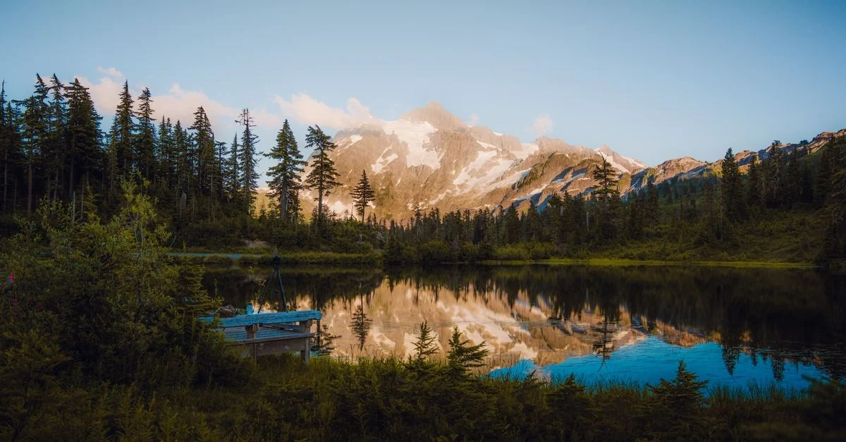 A peaceful mountain lake reflecting trees and snow-capped peaks under a clear sky.