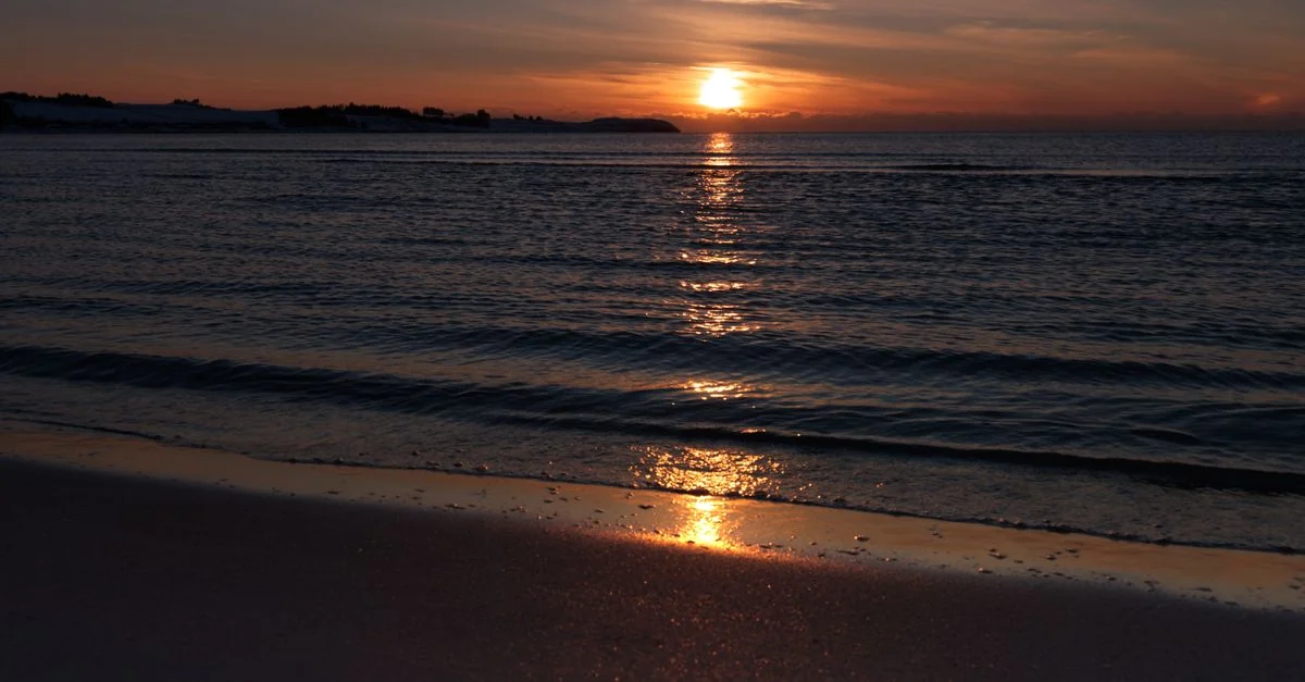 A tranquil sunset over the ocean, with gentle waves and a reflection on the sandy shore.